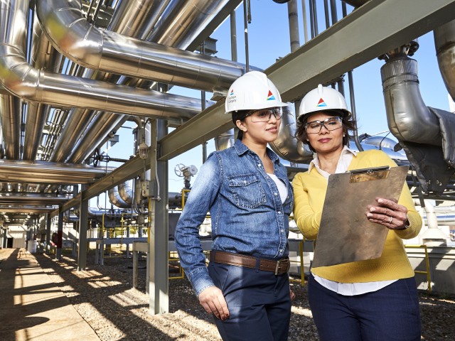 two women wearing hardhats standing under large pipes examining charts