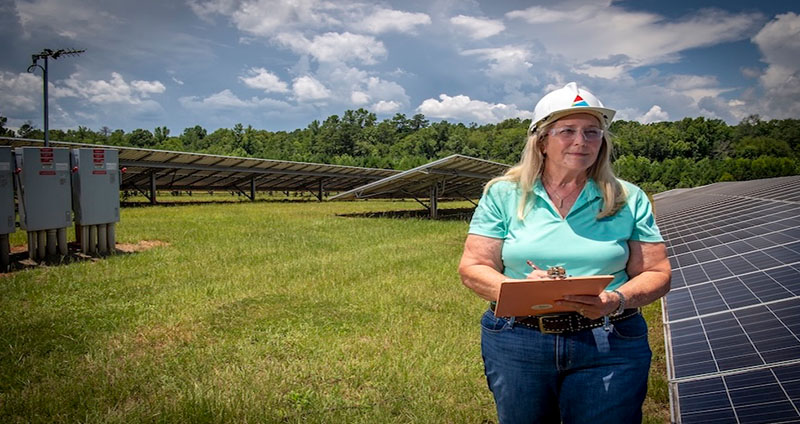 Regina inspects solar panels at a renewable energy site, holding a clipboard and wearing a hard hat and safety gear.