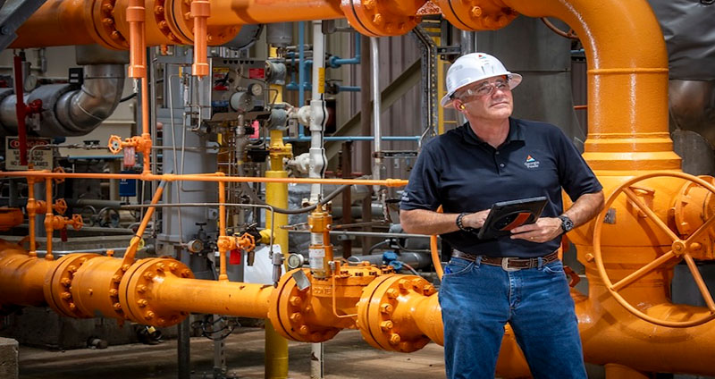 Marcus stands in an industrial setting with large orange pipes, holding a tablet near mechanical equipment and gauges.