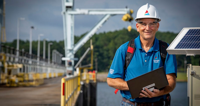 Scott stands near solar equipment at Wallace Dam, wearing a hard hat and holding a laptop, overseeing control systems outdoors.